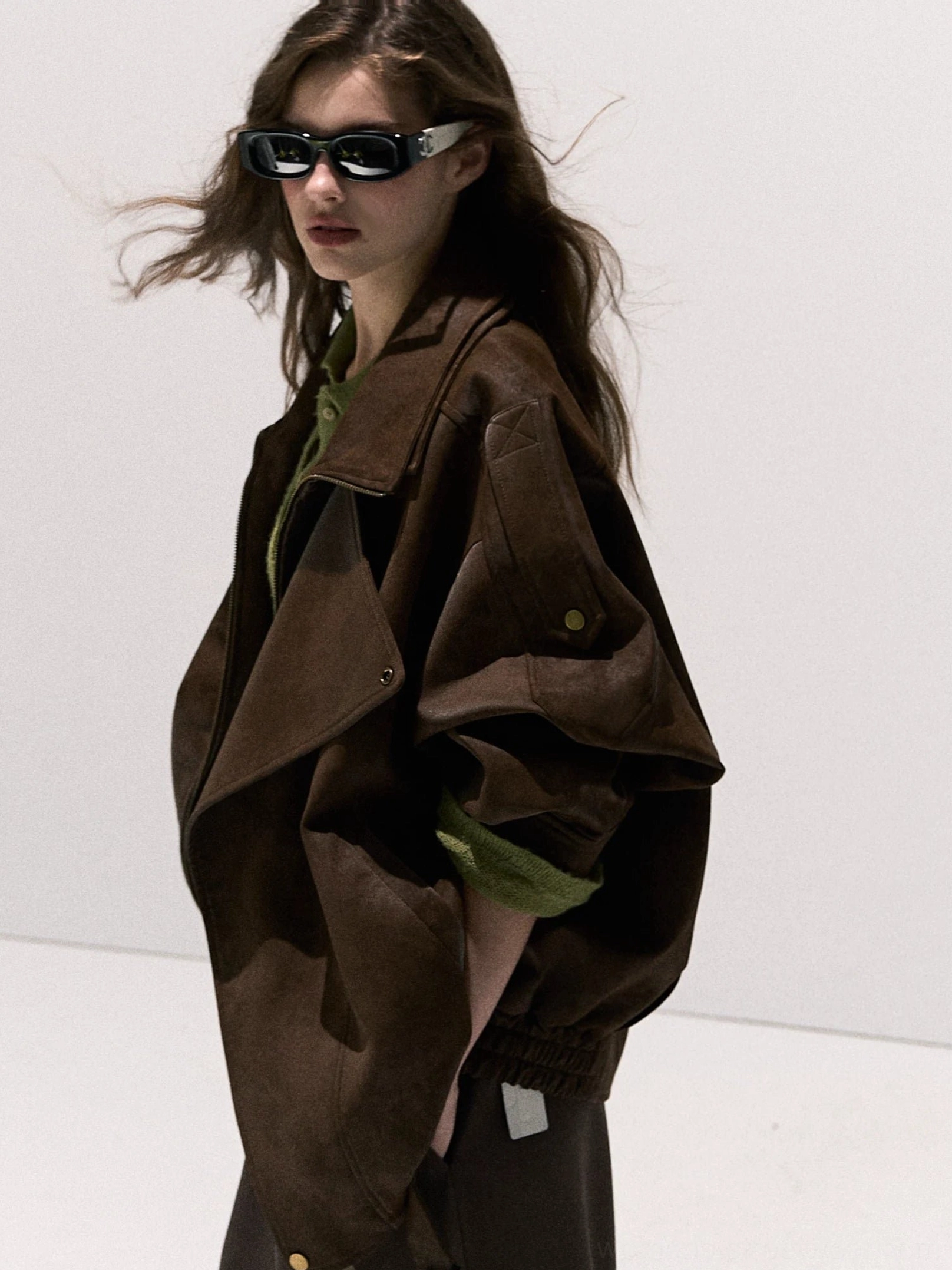 Side view of a brown suede-look oversized moto jacket showing dropped shoulders, snap-button cuffs, elasticated hem, retro texture, green top, and sunglasses