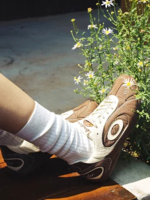 Brown swirl panel retro trainers with dog charm worn with white crew socks among blooming daisy wildflowers