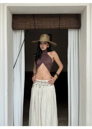 Brown twisted halter neck cut-out crop top styled with a high-waisted maxi skirt, straw hat, and gold jewelry at a resort doorway