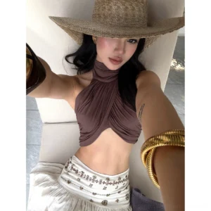 Close-up front view of a brown halter neck crop top with twisted wrap front, gathered fabric, exposed midriff, and gold bangles