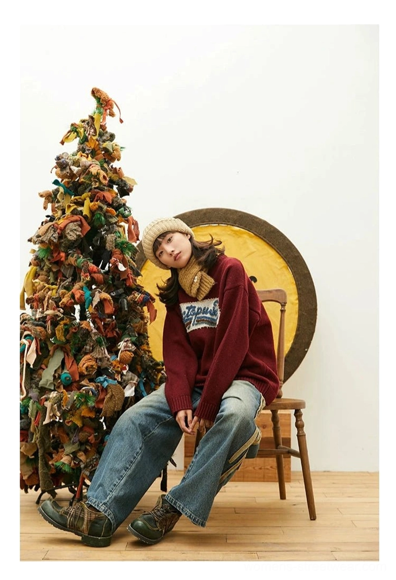Side sitting view of burgundy relaxed crewneck knit sweater styled with vintage washed denim, beige beanie, and long knitted scarf