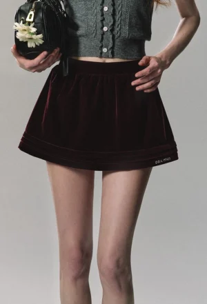 Burgundy velvet pleated mini skirt in an A-line silhouette shown from a low front angle with a floral handbag and streetwear styling