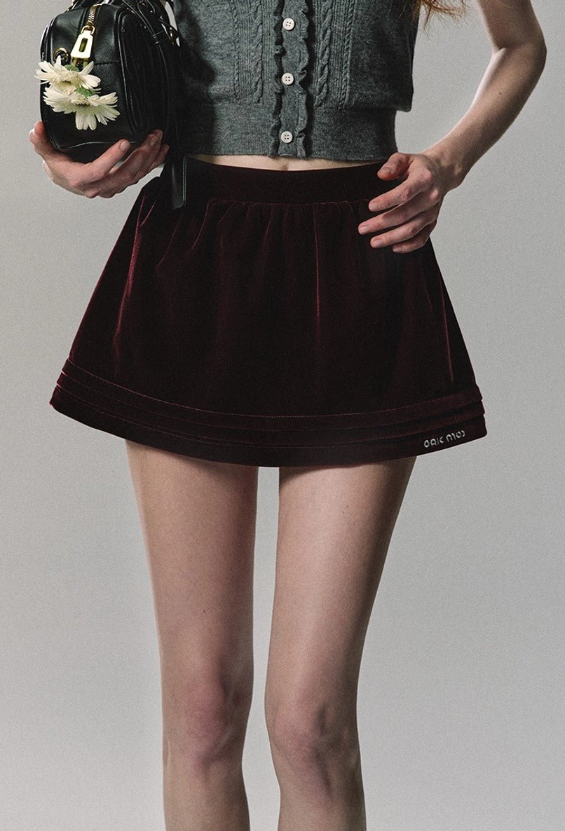 Burgundy velvet pleated mini skirt in an A-line silhouette shown from a low front angle with a floral handbag and streetwear styling