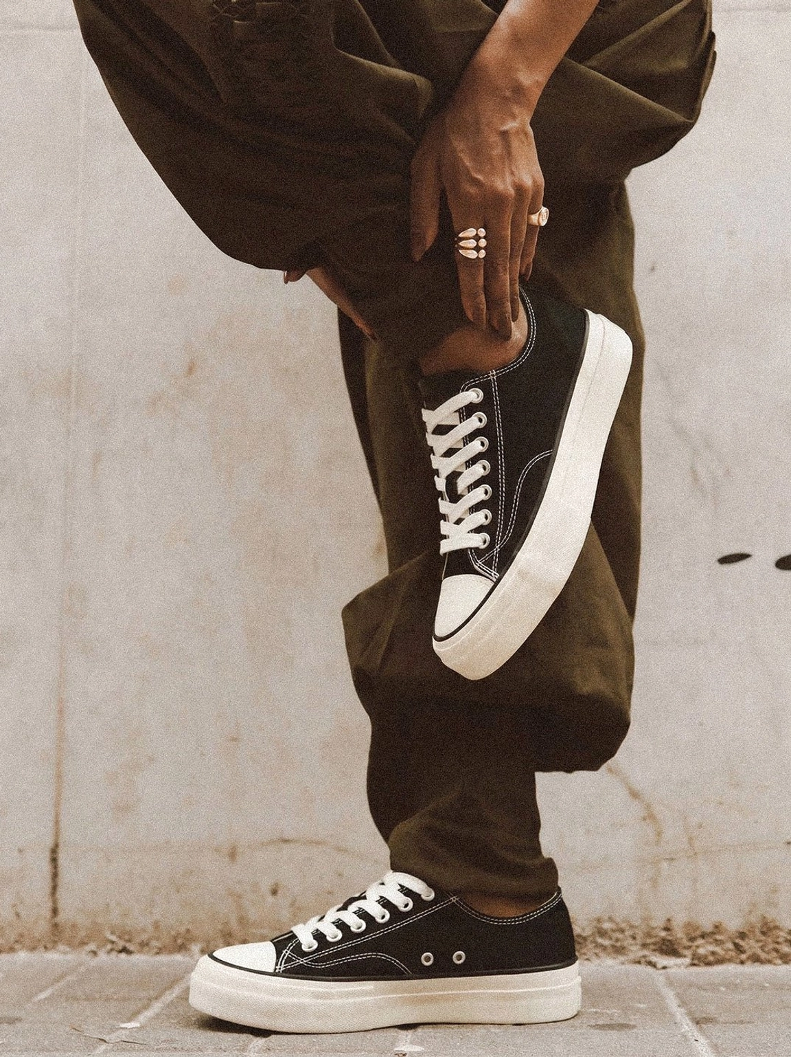 Model in brown cargo pants wearing black and white low-top canvas platform sneakers with cork midsole, leaning against concrete wall casual streetwear