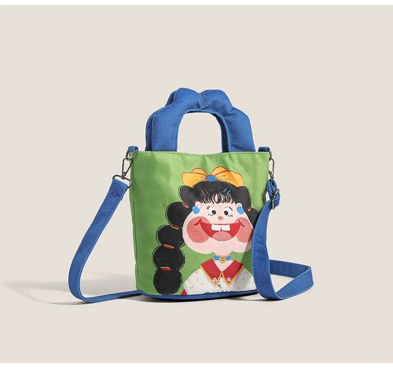 Cartoon girl canvas crossbody tote bag front three-quarter view with green body blue padded handles and playful black-lined girl print streetwear style