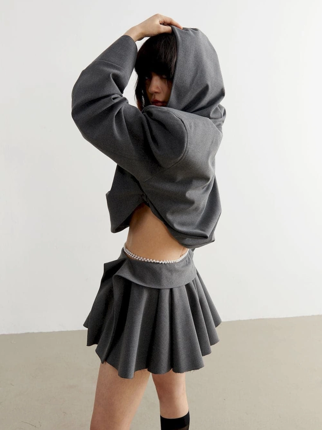 Side back view of charcoal cropped oversized hoodie with loose silhouette and pearl decorated waist styling in a minimalist studio