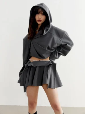 Charcoal cropped oversized hoodie styled with a pleated mini skirt, exposed waist, and pearl waist chain in a minimalist streetwear studio
