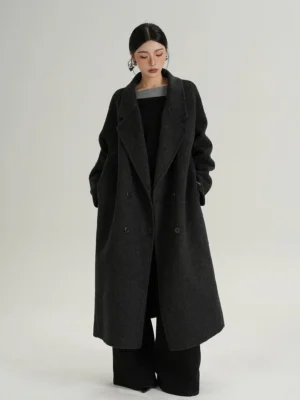 Female model wearing a deep charcoal gray oversized long wool coat with double-breasted matte buttons and side pockets over black wide-leg trousers