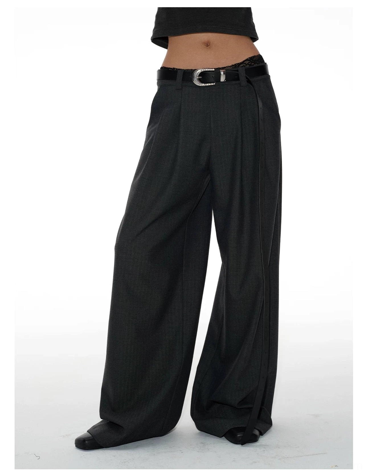 Close-up front view of charcoal gray tailored streetwear trousers showing silver buckle belt, double waist pleats, and smooth suiting fabric