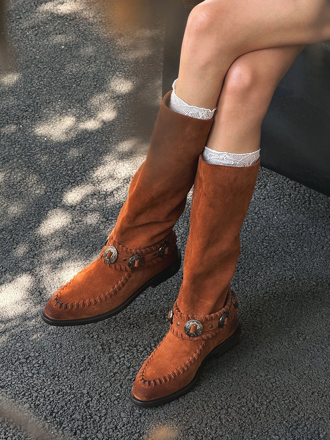 Chestnut brown suede knee-high western boots worn with white ruffled socks in a relaxed seated outdoor streetwear outfit