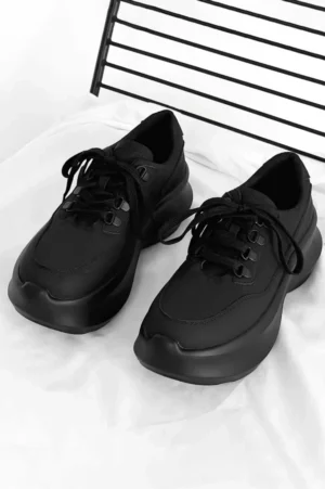 Black chunky platform lace-up sneakers with smooth leather upper shown in a top-down flat lay on white bedding with a metal grille background