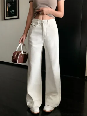 Front low-angle close-up of cream off-white high-waisted wide-leg denim streetwear pants styled with a cropped taupe top white flats and a two-tone handbag