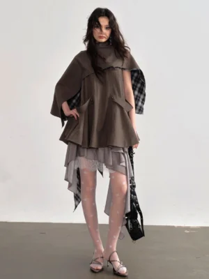 Front view of coffee brown asymmetric cape blouse with high funnel neckline, plaid inner lining, utility pocket details, sheer tights, heels, and black crossbody bag