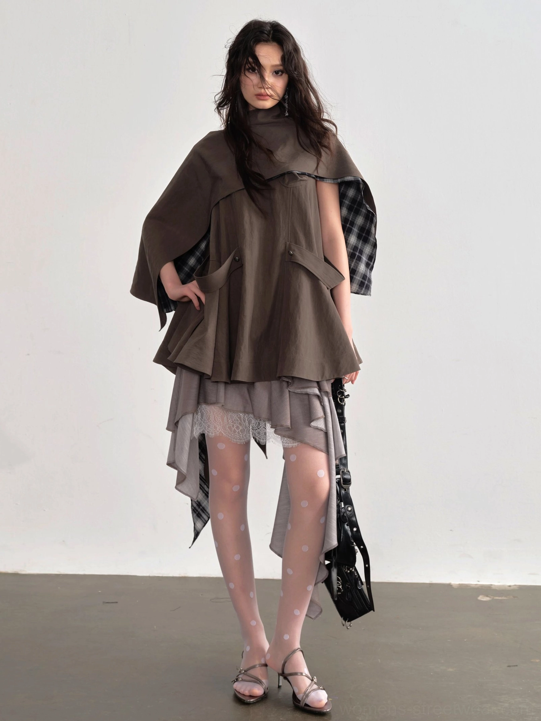 Front view of coffee brown asymmetric cape blouse with high funnel neckline, plaid inner lining, utility pocket details, sheer tights, heels, and black crossbody bag