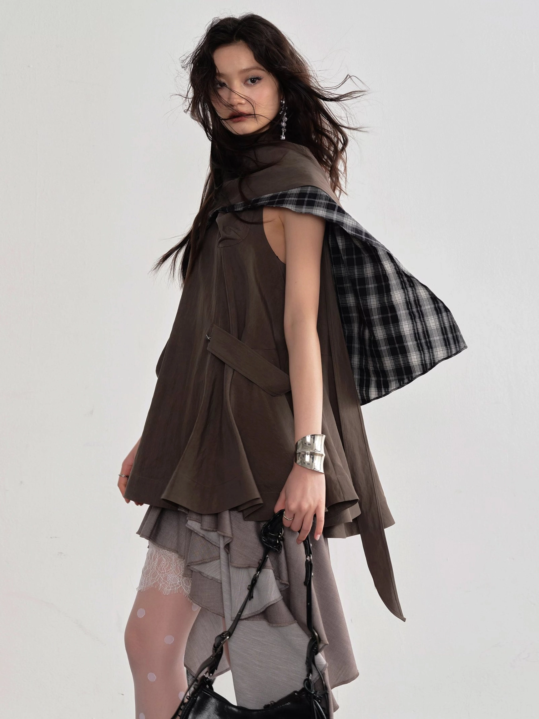 Coffee brown asymmetric cape layer blouse shown from a left three quarter angle with plaid lining, irregular hem, sheer polka dot tights, strappy heels, and black shoulder bag