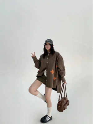 Front three-quarter view of a loose coffee green embroidered button shirt styled with green shorts, baseball cap, and brown handbag