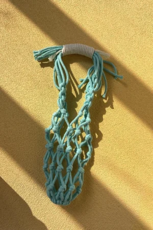 Cyan blue cotton macrame water bottle carrier shown from above on a warm yellow tabletop with hand-knotted net pocket, wrapped handle, and tassel details
