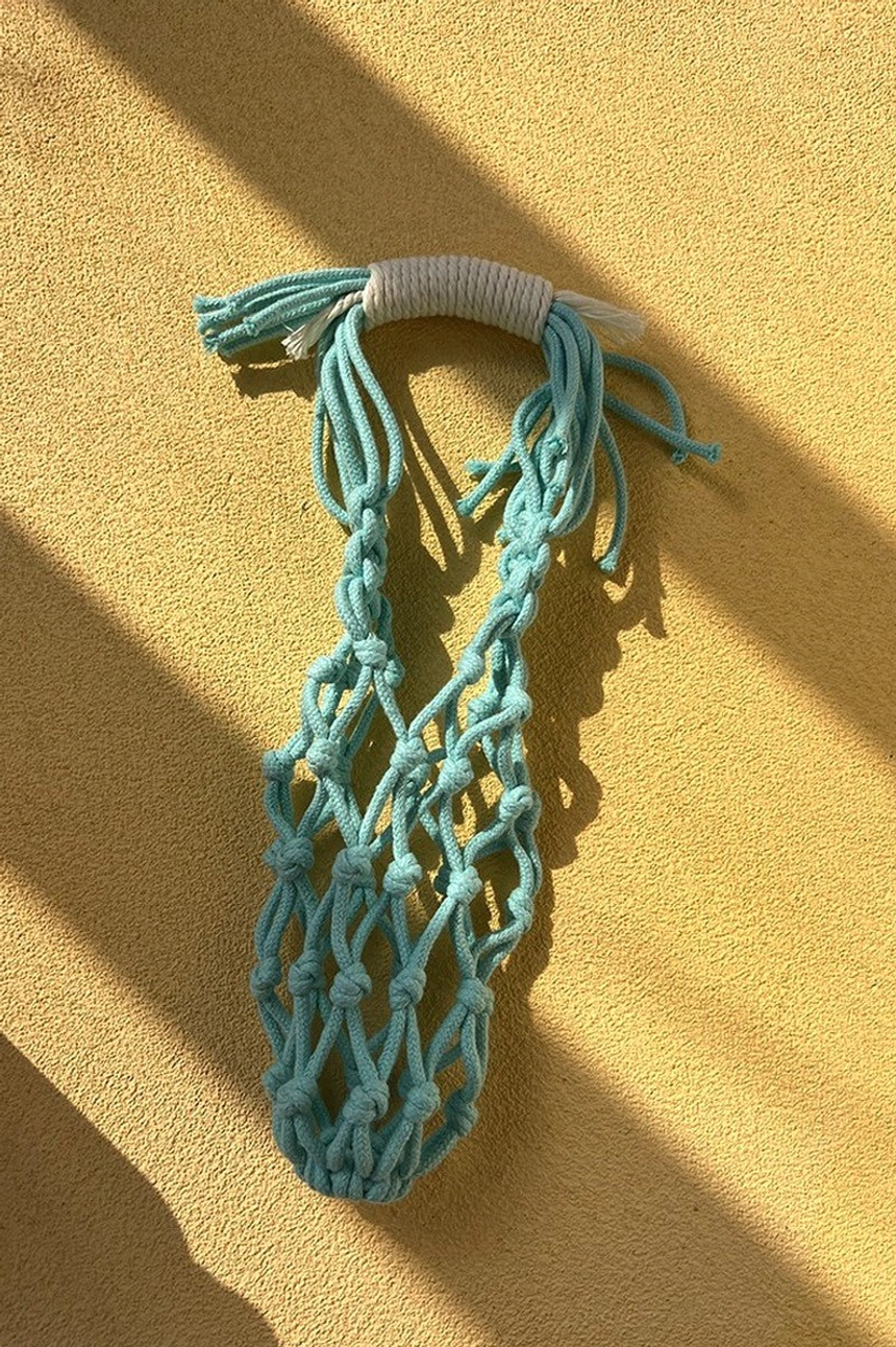Cyan blue cotton macrame water bottle carrier shown from above on a warm yellow tabletop with hand-knotted net pocket, wrapped handle, and tassel details