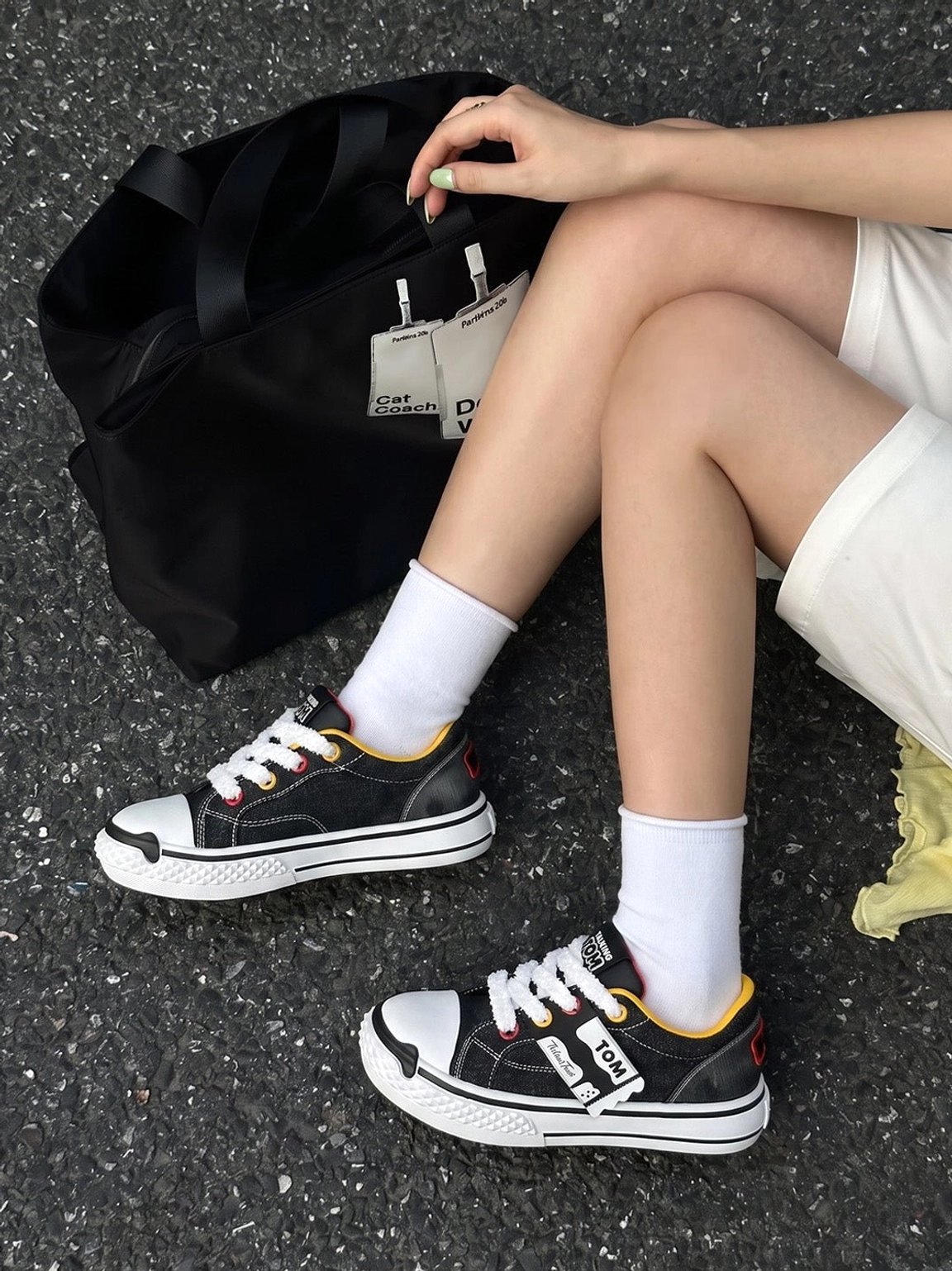 Contrast graphic chunky sneakers worn with white shorts and socks in a streetwear flat lay on asphalt beside a black tote bag