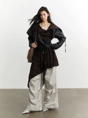 Front three-quarter view of a black convertible faux leather jacket with asymmetric dark plaid layered panel styled with oversized beige cargo pants