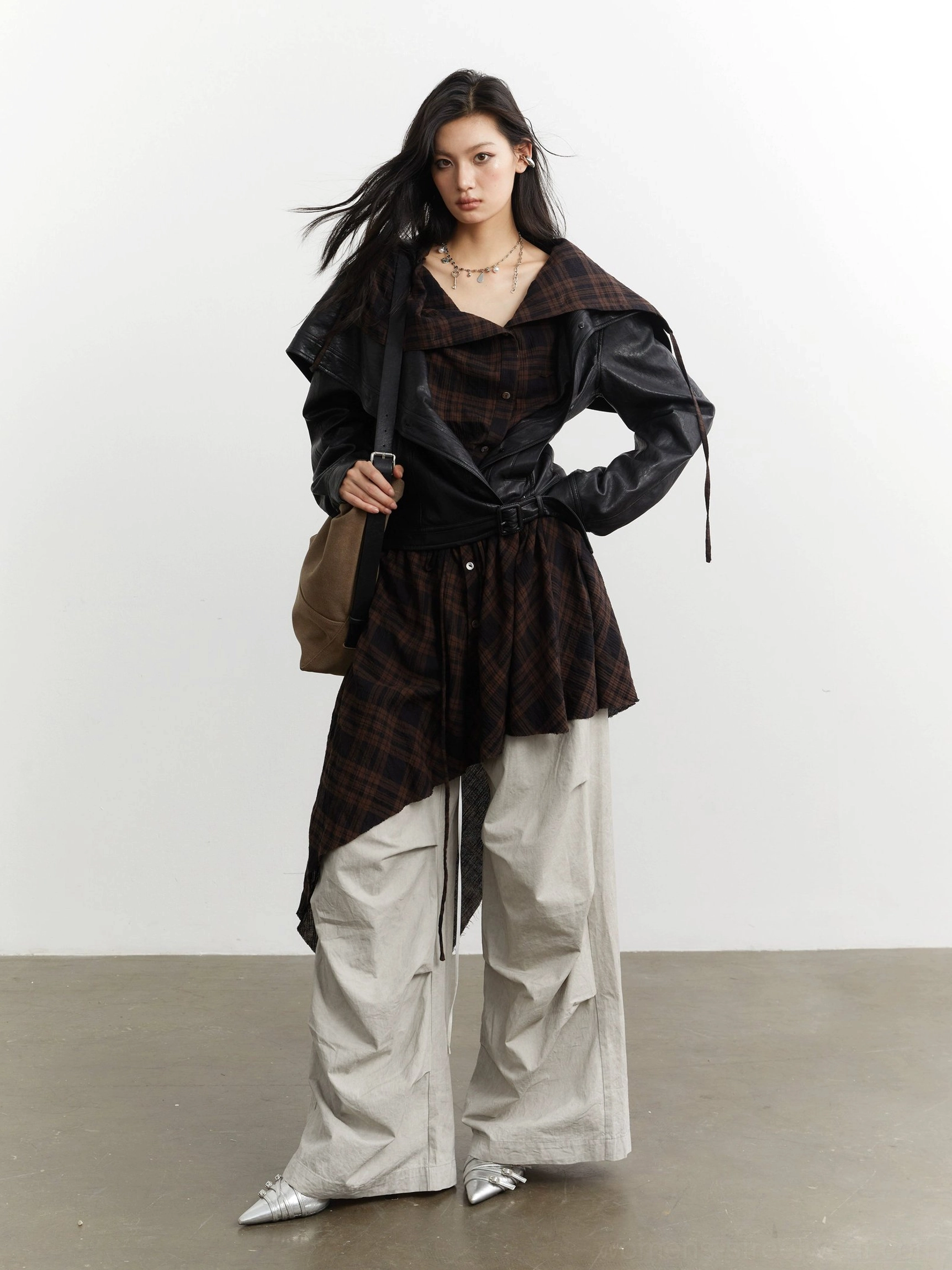 Front three-quarter view of a black convertible faux leather jacket with asymmetric dark plaid layered panel styled with oversized beige cargo pants