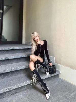 Model wearing black corset lace-up knee-high stiletto boots with an all-black streetwear outfit at an outdoor stairwell