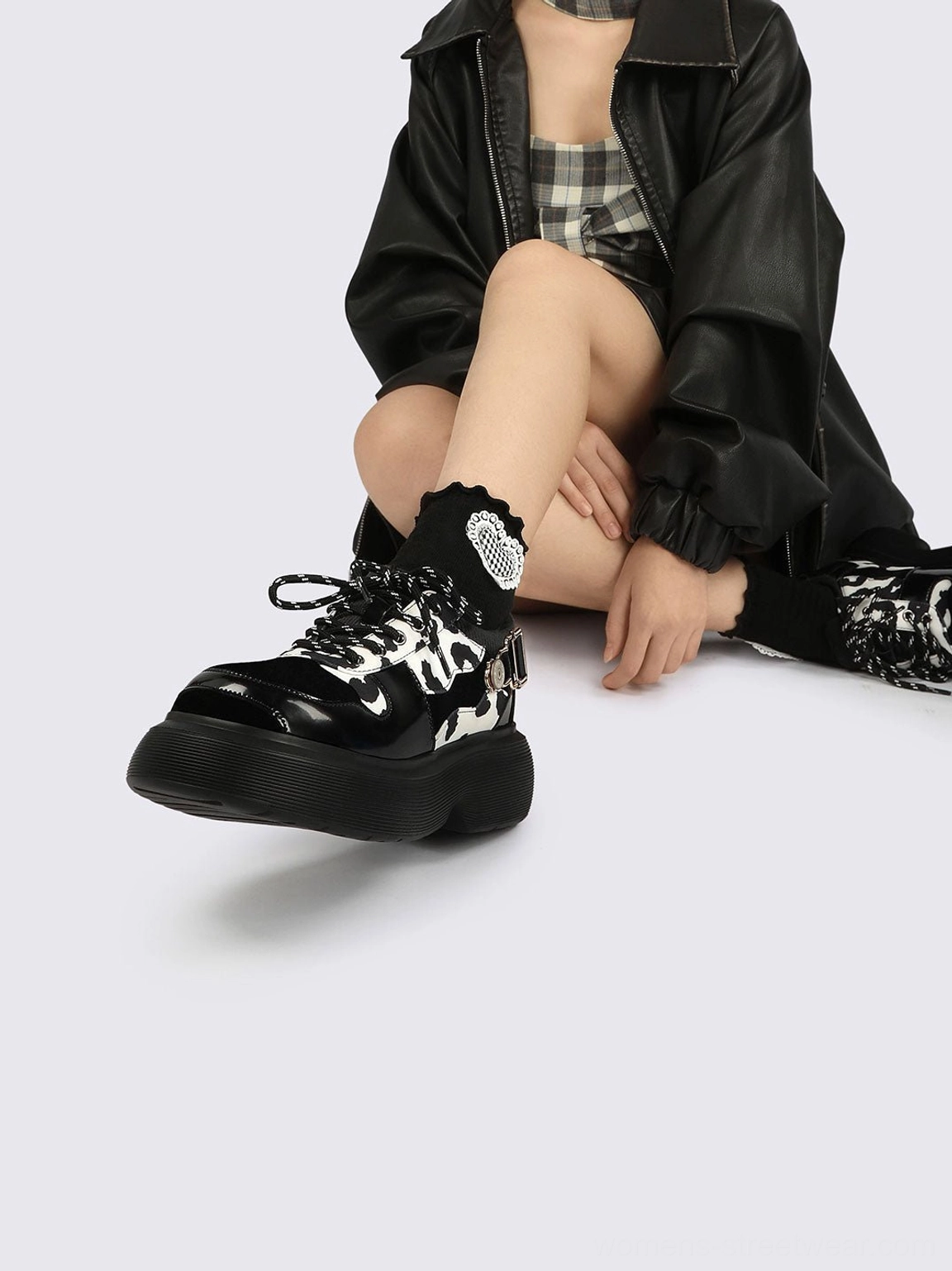 Low-angle side view of cow print platform shoes with lace-up fronts and chunky soles worn with a black leather jacket and plaid outfit