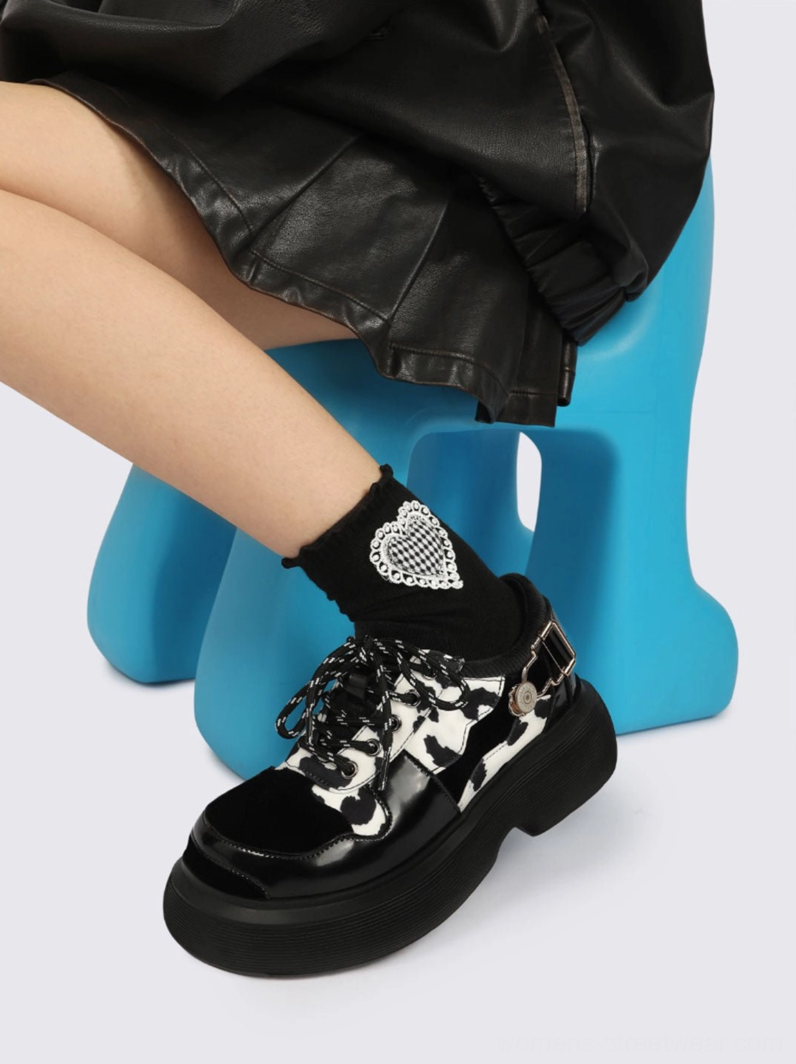 Low-angle side view of cow print platform lace-up Mary Jane shoes with decorative buckle straps, black skirt, and heart-patterned socks