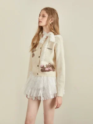 Left three-quarter view of a cream beige teddy bear print cropped cardigan with fluffy jacquard knit details, front buttons, embroidered chest lettering, lace scarf, and white ruffle skirt
