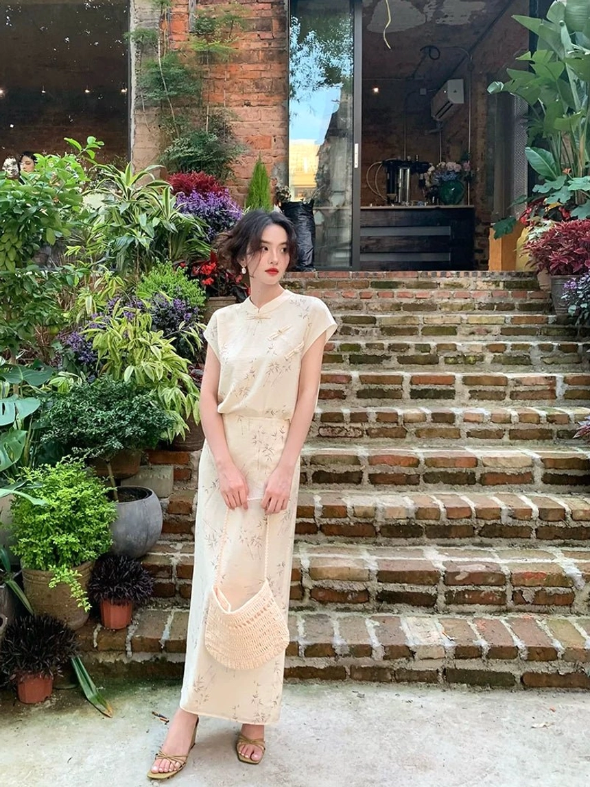Full-body view of a woman wearing a cream floral cheongsam-style blouse and long skirt streetwear set with a mandarin collar, cap sleeves, high-waisted flowing silhouette, and subtle botanical print