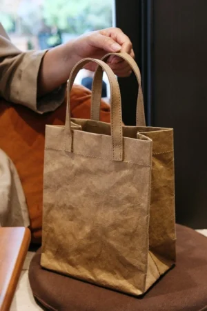 Brown crinkled kraft paper effect cotton tote bag with soft double top handles displayed on a stool in a cozy interior