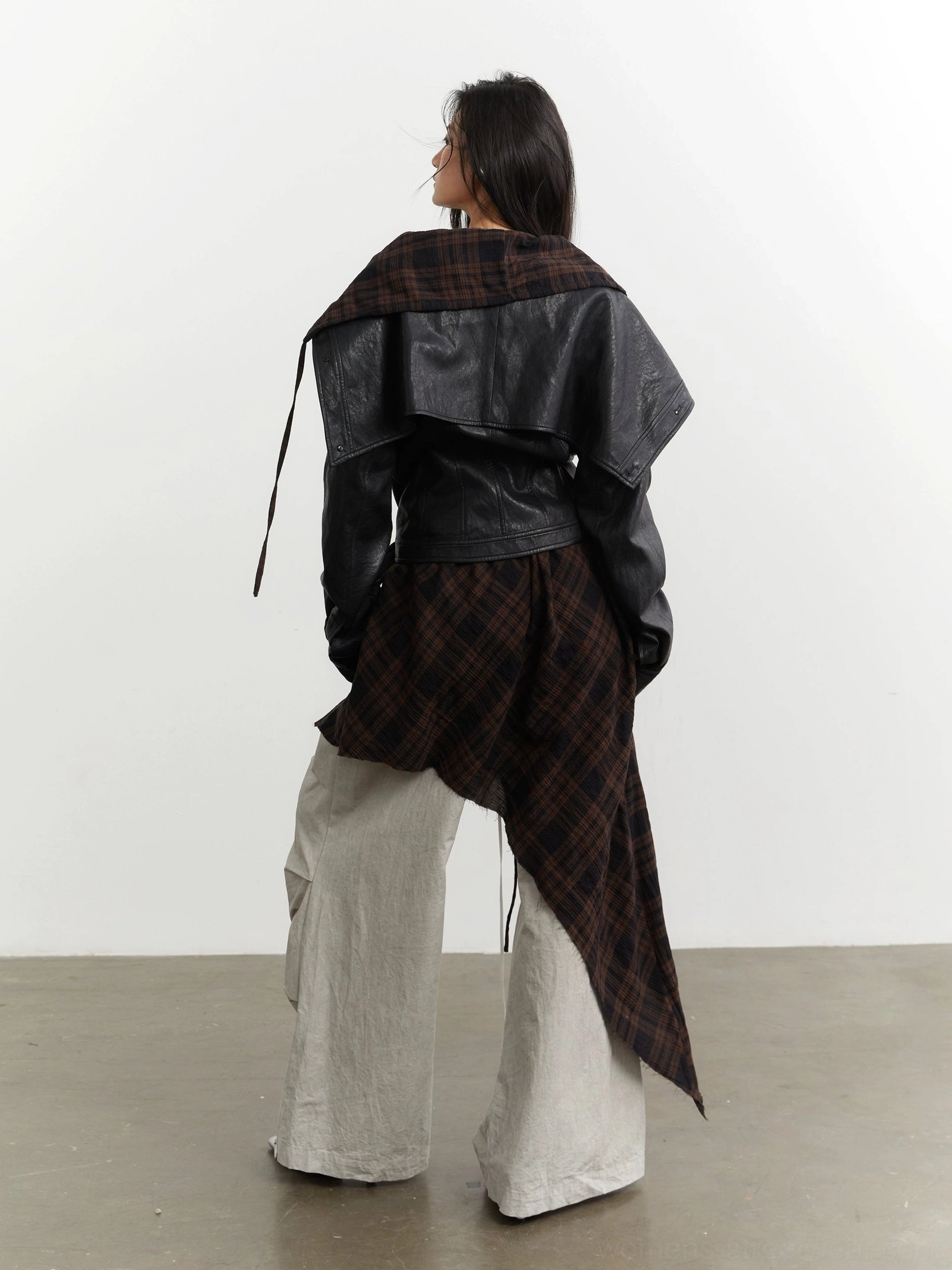 Back three-quarter view of a cropped black faux leather jacket with long asymmetric dark tartan wrap skirt panel and adjustable side ties