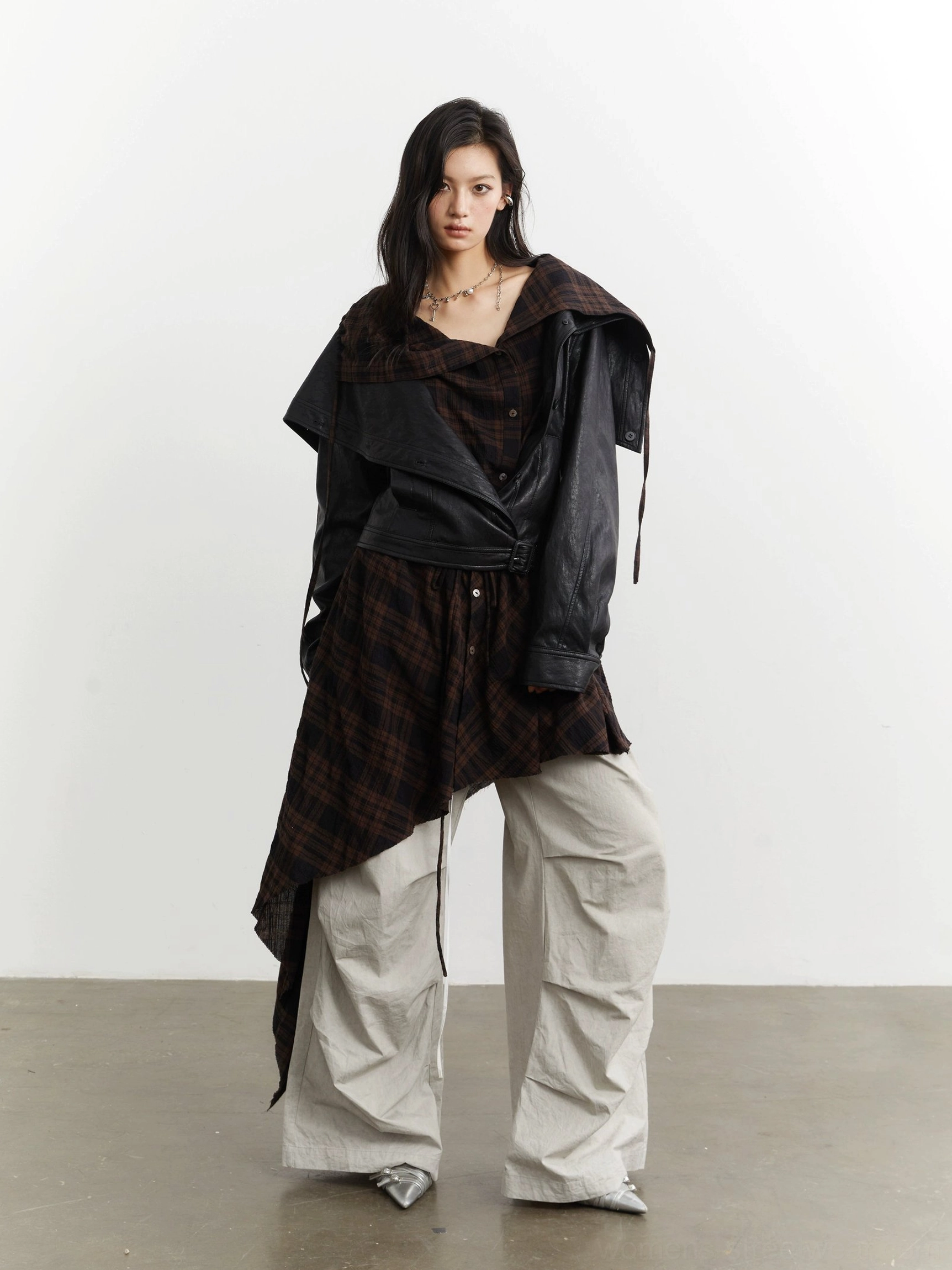 Front three-quarter streetwear styling of a cropped black leather-look jacket with tied asymmetric plaid underlayer and loose cargo trousers