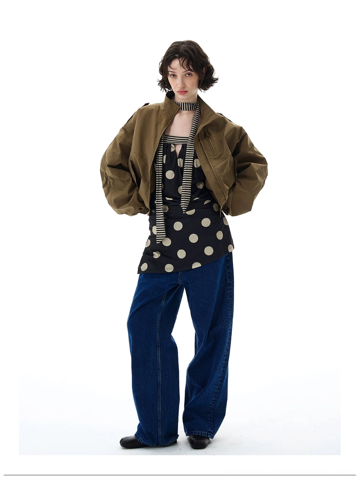Female model wearing a cropped olive green utility bomber jacket with high stand collar, relaxed sleeves, and wide-leg denim in a white studio