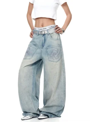 Front full-body view of ice-blue oversized wide-leg Y2K streetwear jeans with rhinestone starfall details, embroidered script patch pockets, silver belt, and cropped white tee