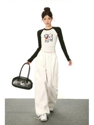 Female model walking in a cartoon Dalmatian print cropped long sleeve tee with black raglan sleeves, white cargo pants, sneakers, and black shoulder bag