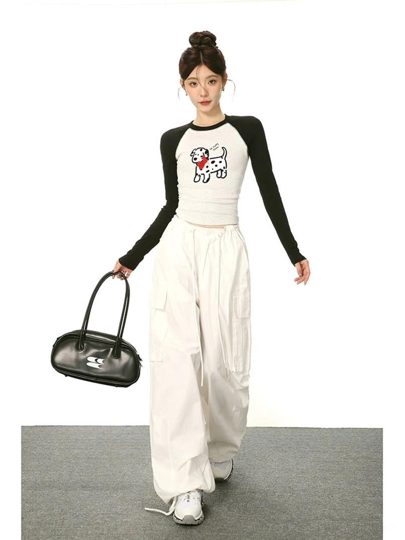 Female model walking in a cartoon Dalmatian print cropped long sleeve tee with black raglan sleeves, white cargo pants, sneakers, and black shoulder bag