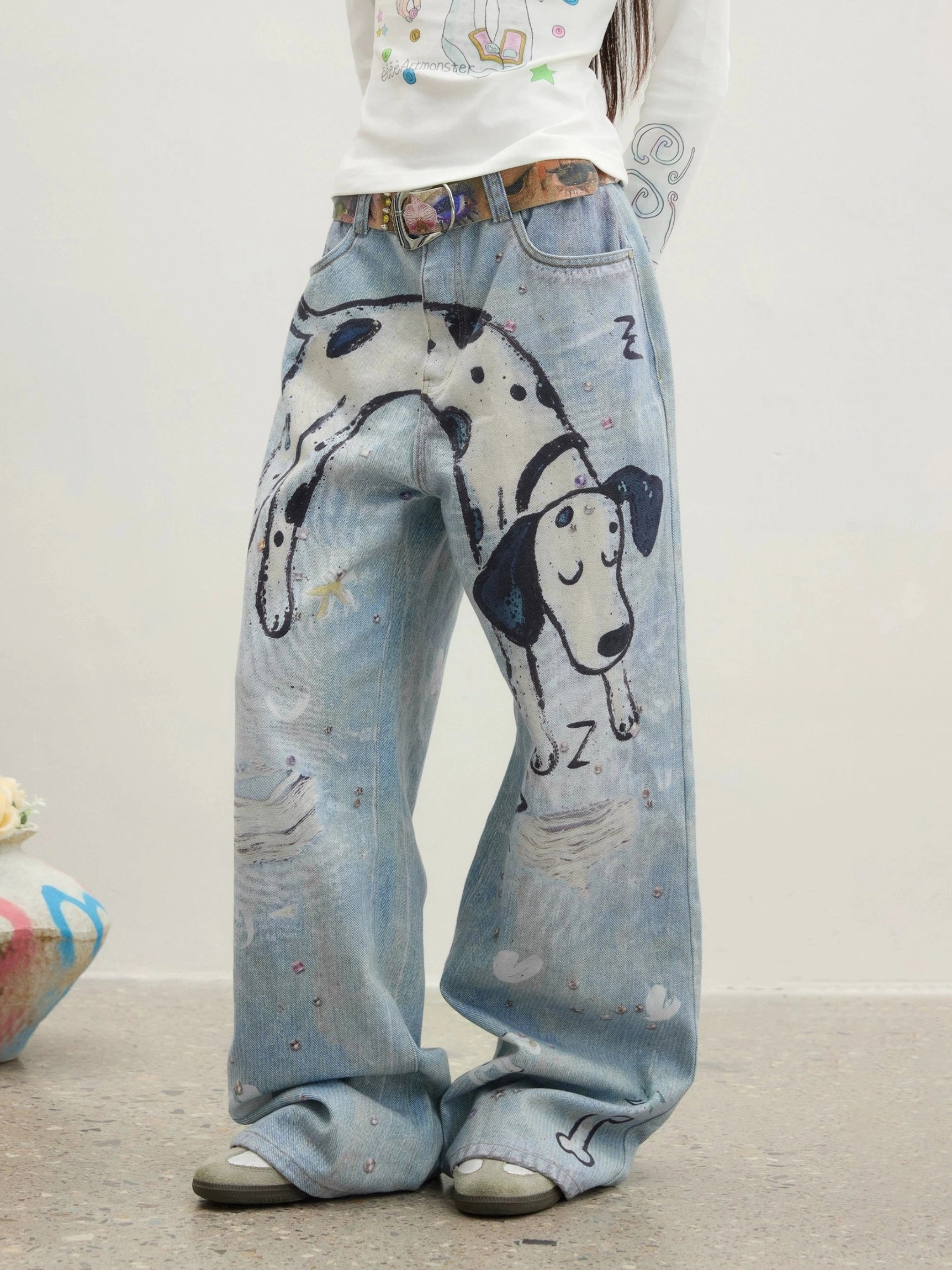 Close-up view of washed blue straight-leg jeans waistband and upper legs showing belt loops, colorful belt styling, sleeping dalmatian graphic, and decorative denim speckles