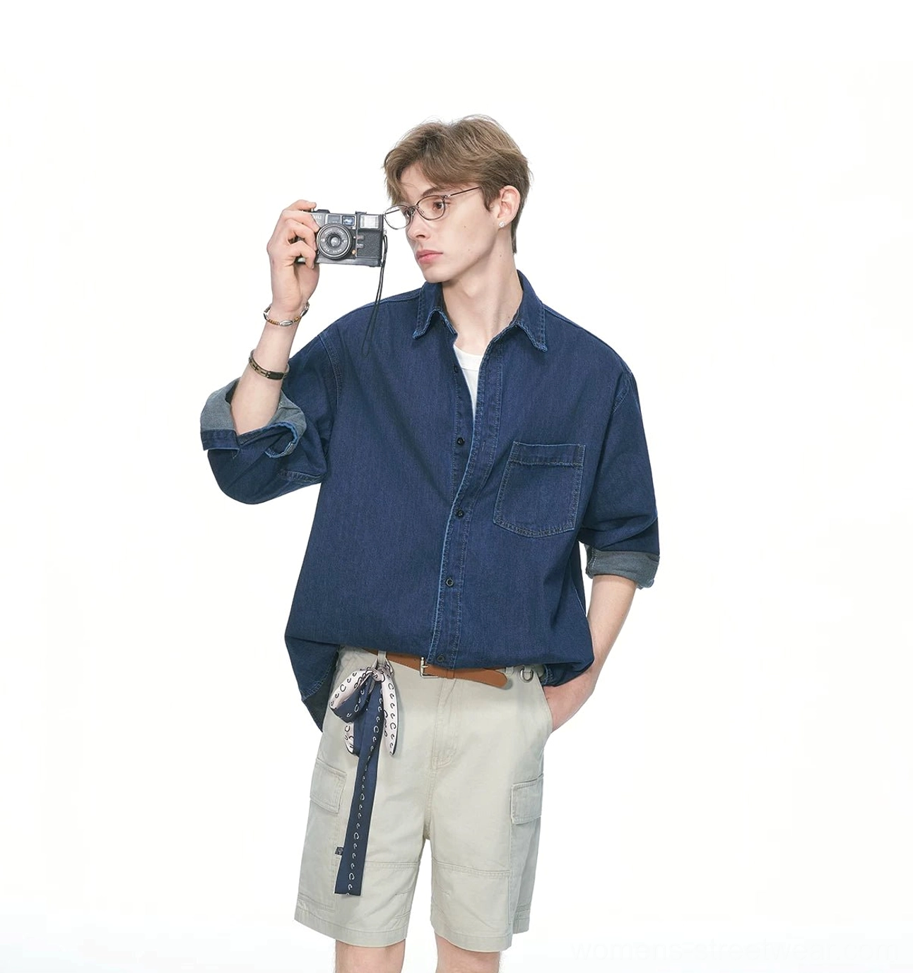 Male model wearing a dark blue oversized denim shirt with rolled cuffs, khaki cargo shorts, bandana belt, and vintage film camera