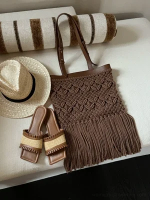 Dark brown macrame fringe openwork tote bag styled on a light fabric sofa with a straw hat and woven slide sandals