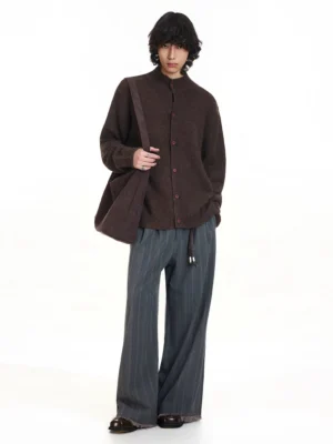 Full body view of a dark brown oversized knitted cardigan styled with striped gray wide-leg trousers and a brown shoulder tote bag