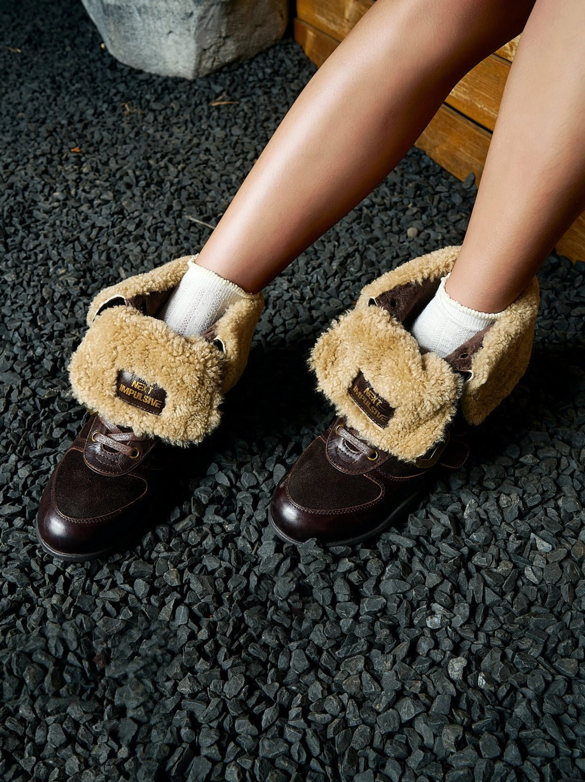 Dark brown shearling leather ankle boot styled with white crew socks in an outdoor gravel setting