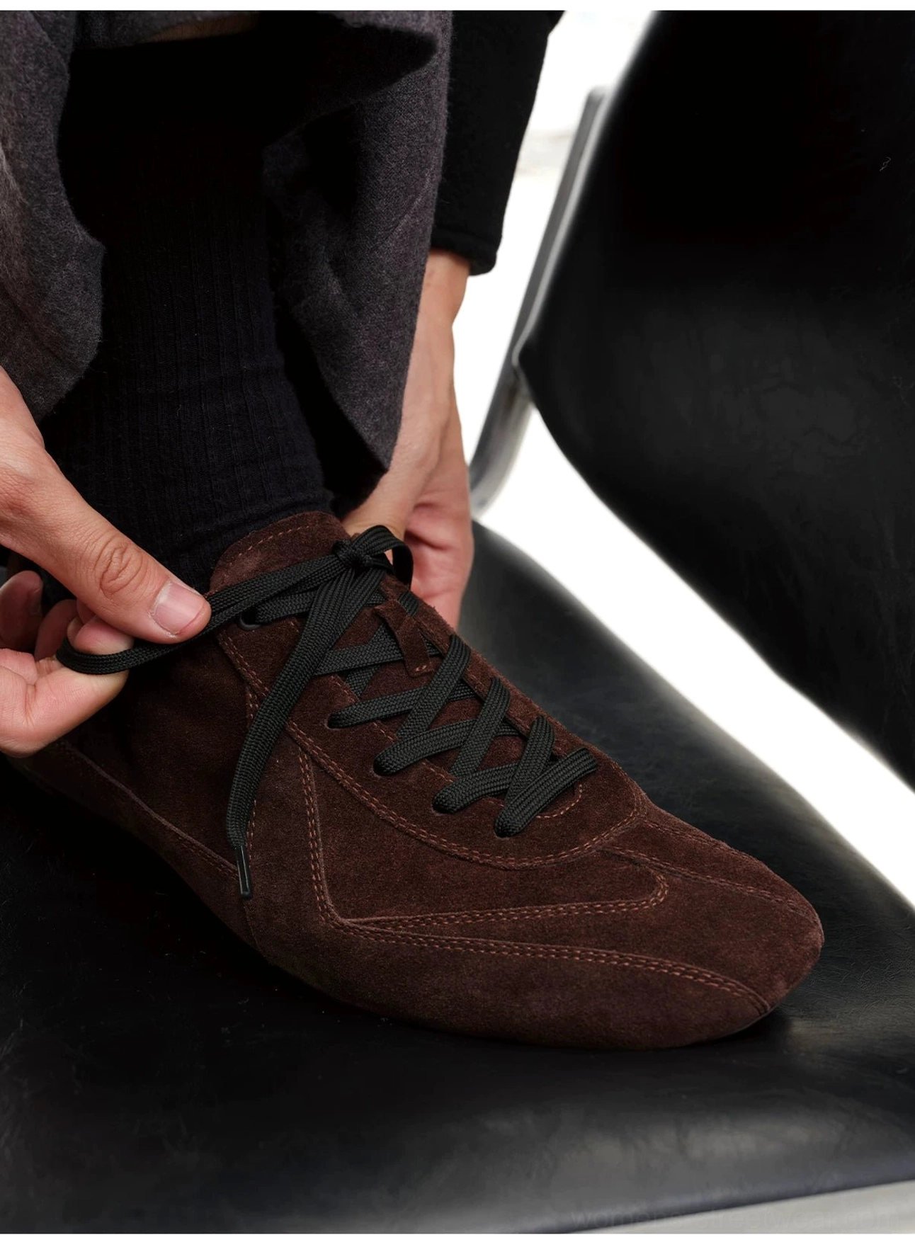 Close-up of dark brown suede minimalist sneakers being tightened by hand, featuring flat black laces and a low-top design