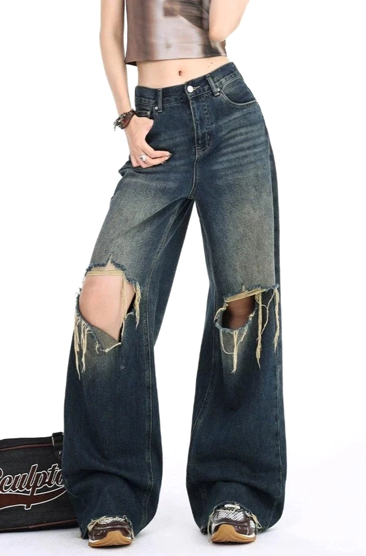 Front three-quarter view of dark gradient distressed wide leg ripped jeans with large frayed knees, high-rise waist, relaxed denim fit, cropped top, and vintage streetwear accessories