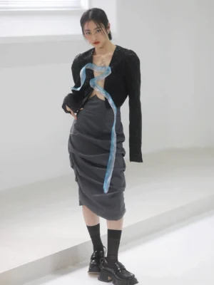 Front full-body view of a woman wearing a dark gray asymmetric ruched tulip midi skirt styled with a cropped black textured jacket, blue organza ribbon, black socks, and platform loafers in a minimalist studio