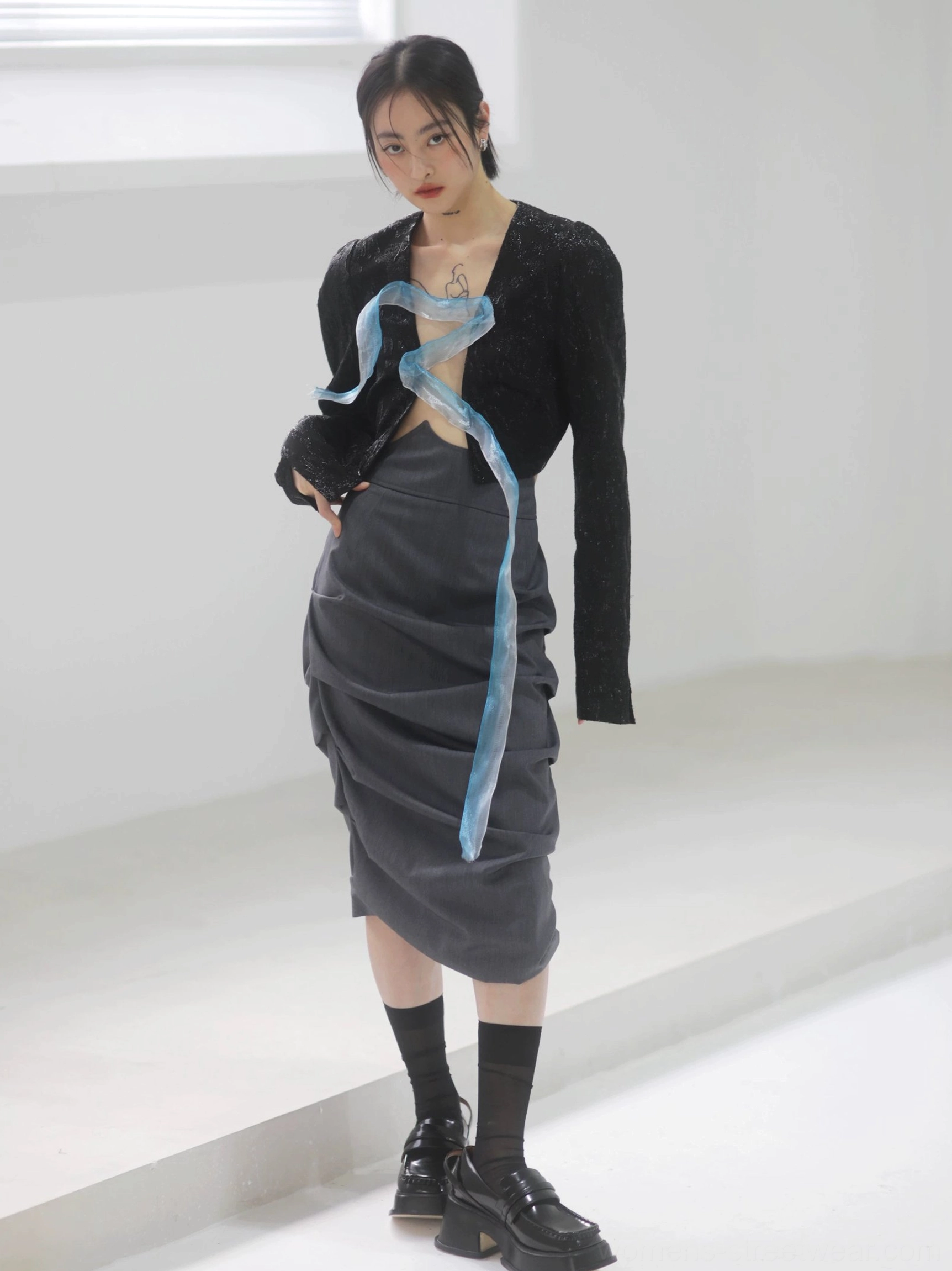 Front full-body view of a woman wearing a dark gray asymmetric ruched tulip midi skirt styled with a cropped black textured jacket, blue organza ribbon, black socks, and platform loafers in a minimalist studio