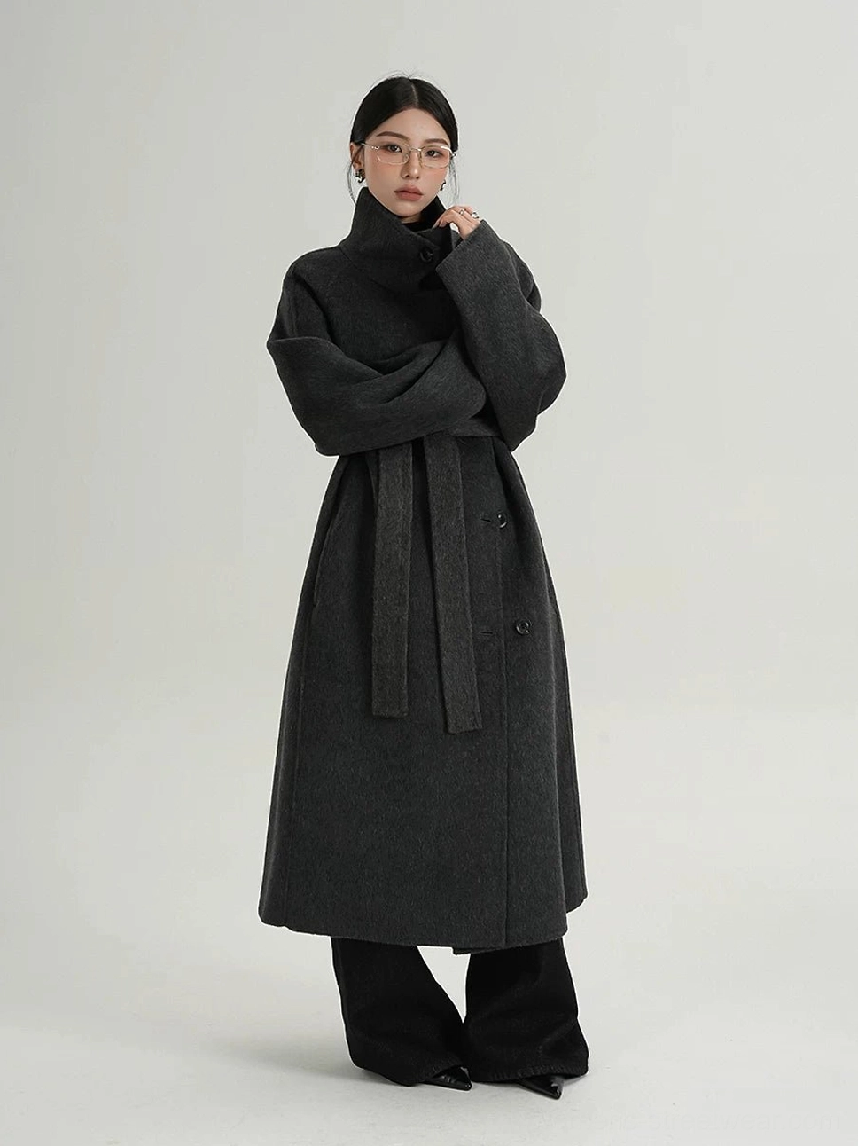 Dark gray oversized funnel neck wool coat styled with a detachable waist belt, buttoned front, long sleeves, and black wide-leg pants