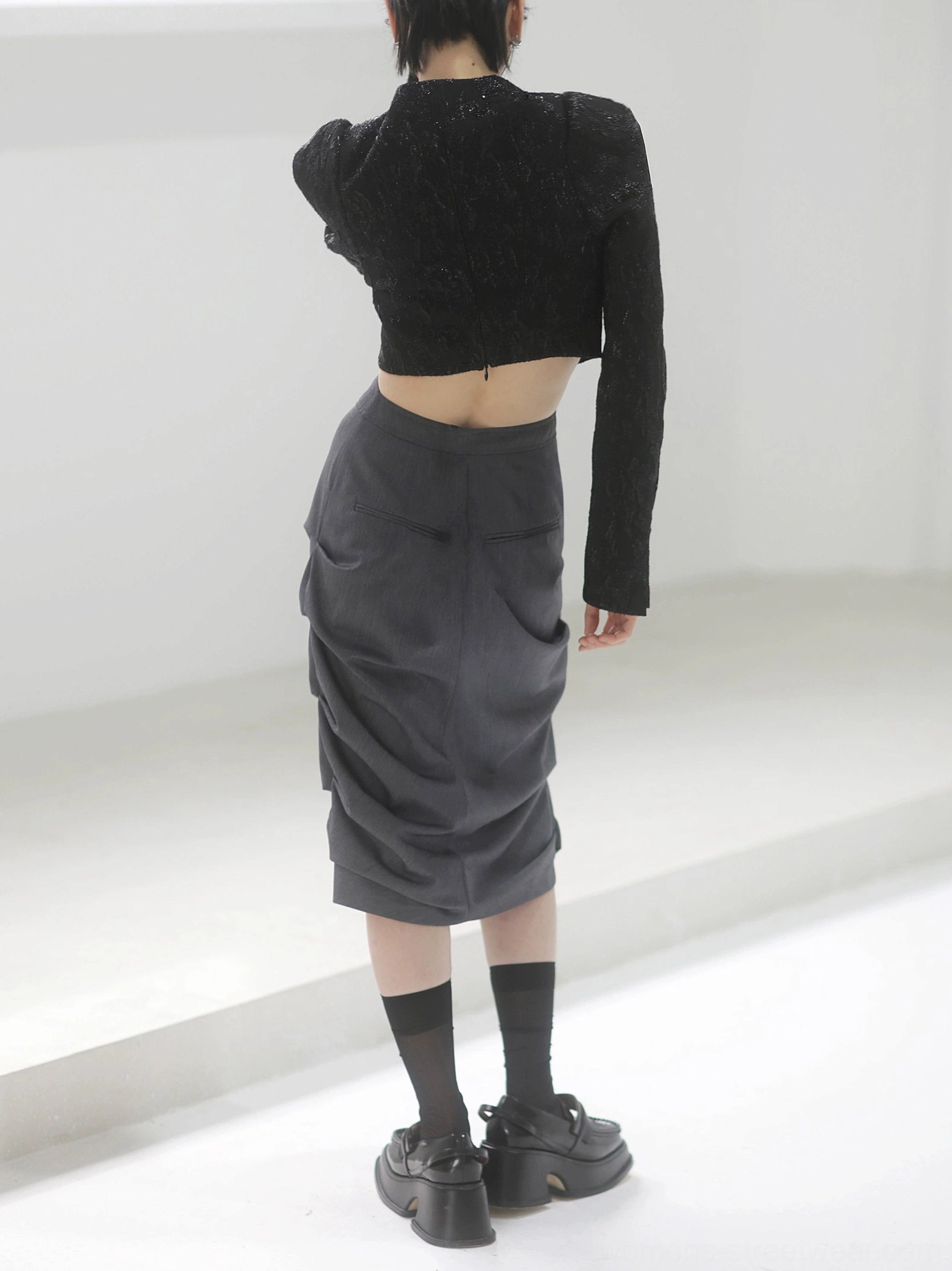 Back full-body view of a dark gray gathered midi skirt with a tapered tulip silhouette styled with a cropped black open-back jacket and chunky platform shoes