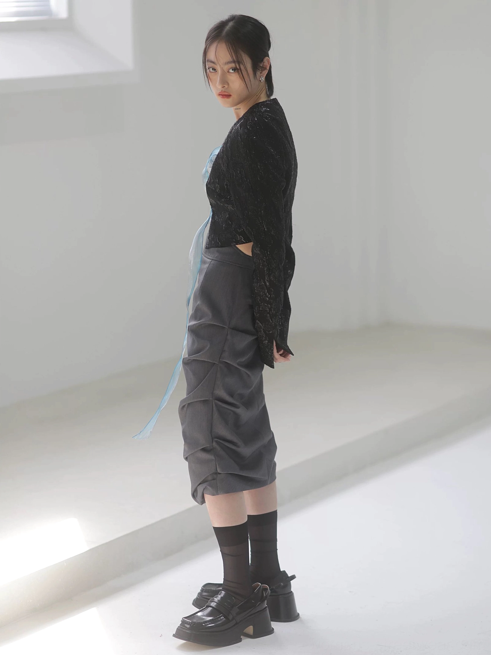 Three-quarter side view of a dark gray high-waisted ruched midi skirt showing the asymmetric side drape and sculptural folds with a cropped black jacket and blue sheer ribbon