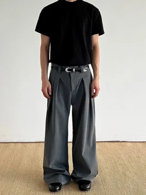 Dark gray pleated wide-leg streetwear trousers shown from the front on a model wearing a black T-shirt, featuring relaxed drape, double front pleats, and full-length tailored legs